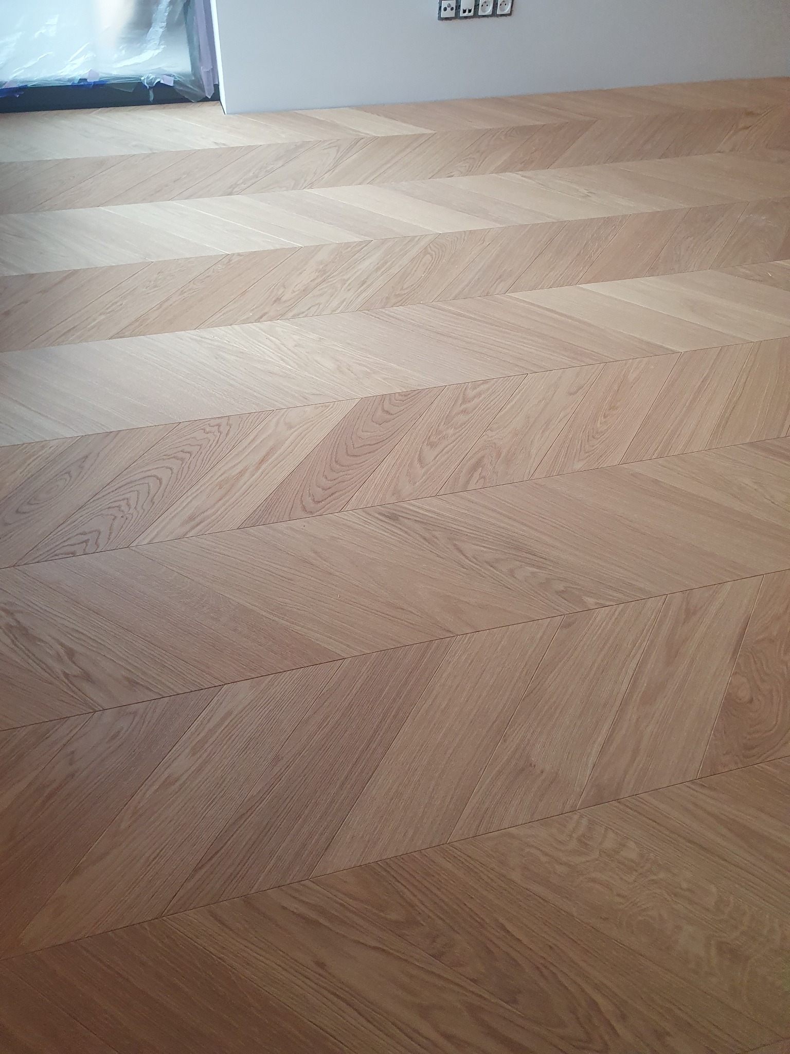 Jasny parkiet ułożony we wzór jodełki francuskiej, widoczne gniazdka elektryczne na ścianie i folia ochronna na ramie okna.