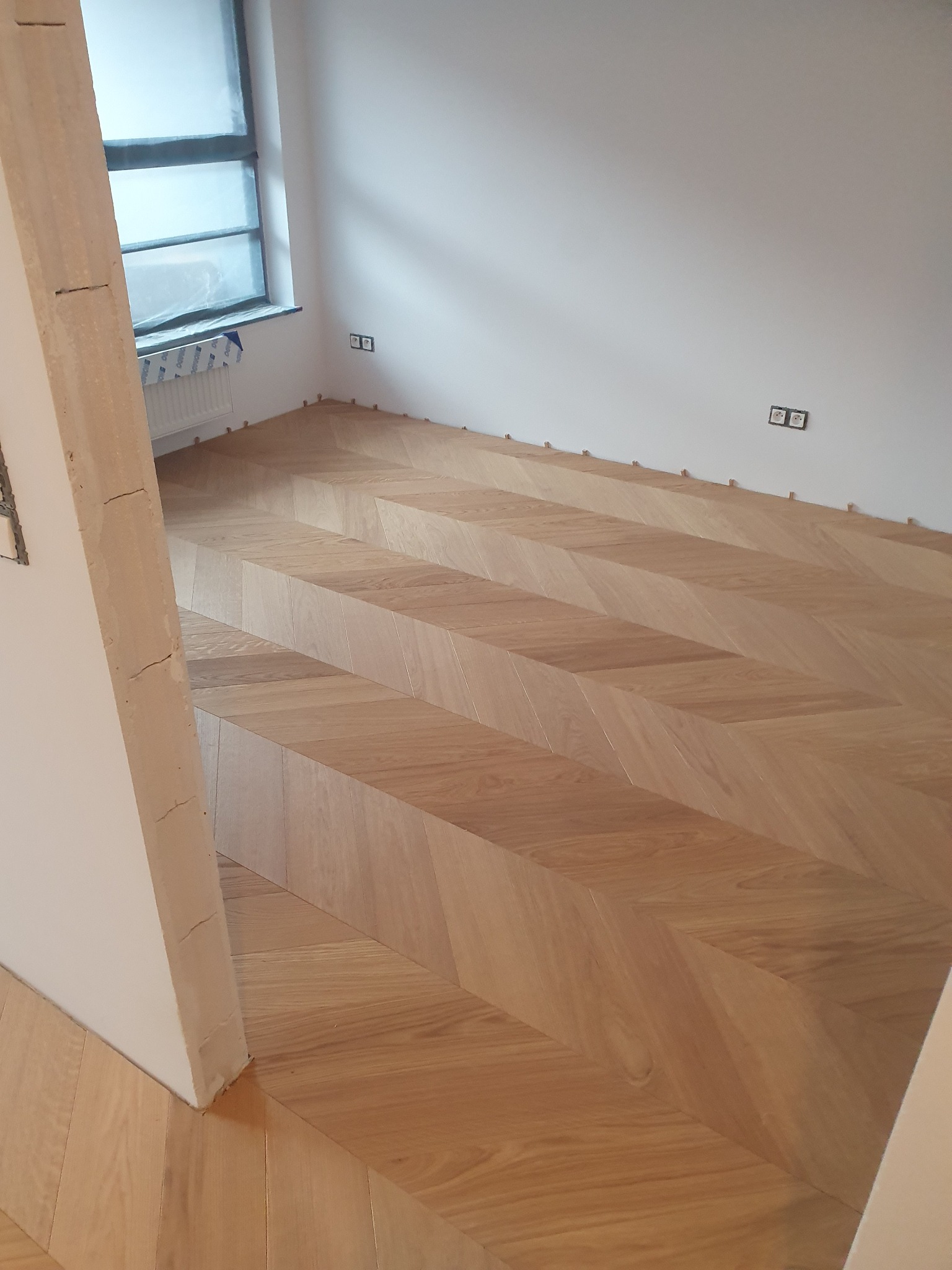 Układanie parkietu w jodełkę francuską, widoczne kliny dystansowe przy ścianie, surowy filar w tle i okno zabezpieczone folią.
