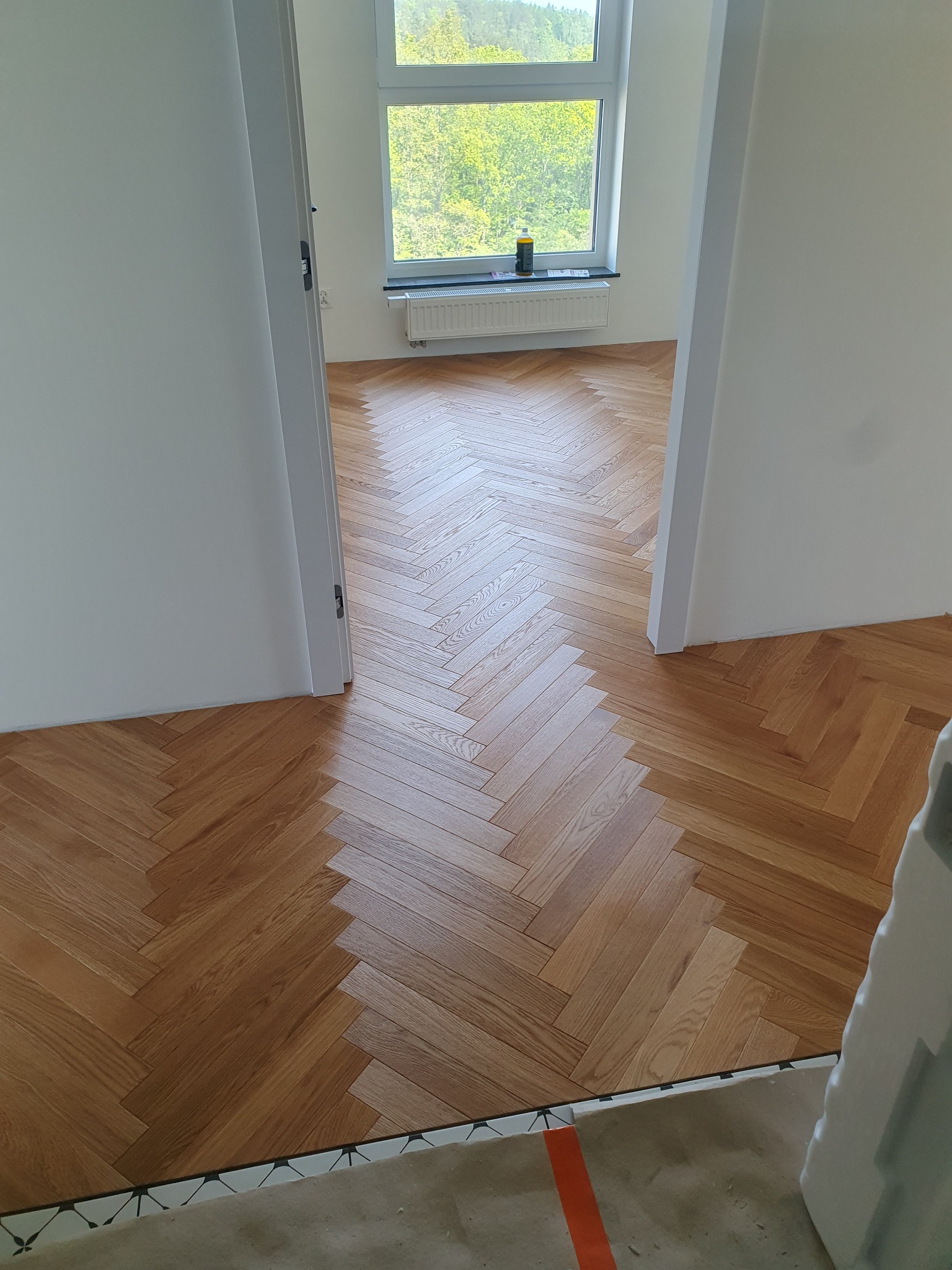 Ułożony parkiet w jodełkę w jasnym pomieszczeniu z białymi ścianami, widok przez otwarte drzwi na dalszą część podłogi i okno z butelką na parapecie.