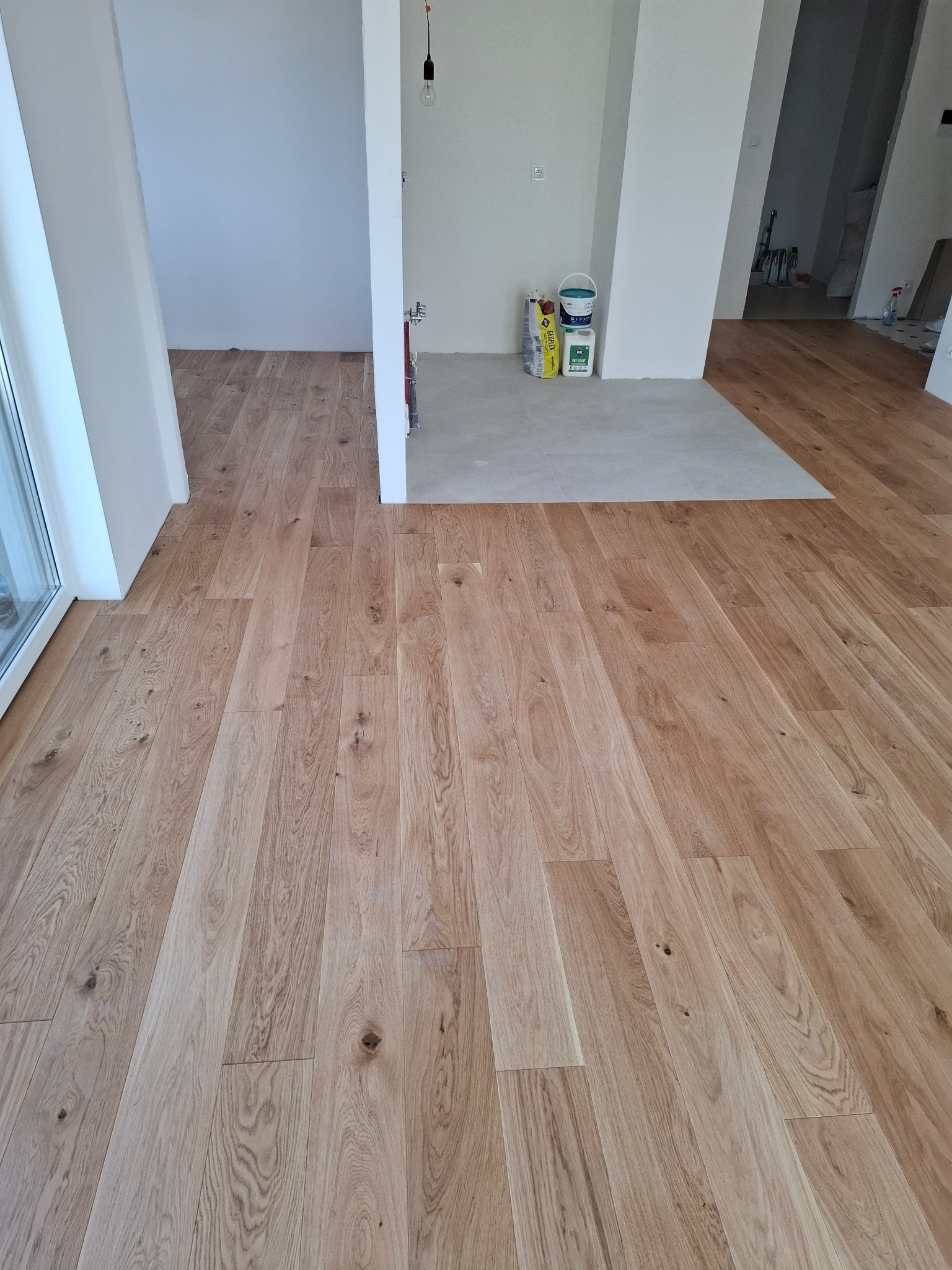 Jasny, dębowy parkiet ułożony w nowym, jasnym wnętrzu. Widoczne puszki z farbą i worki z zaprawą w tle, sugerujące świeżo zakończone prace wykończeniowe.