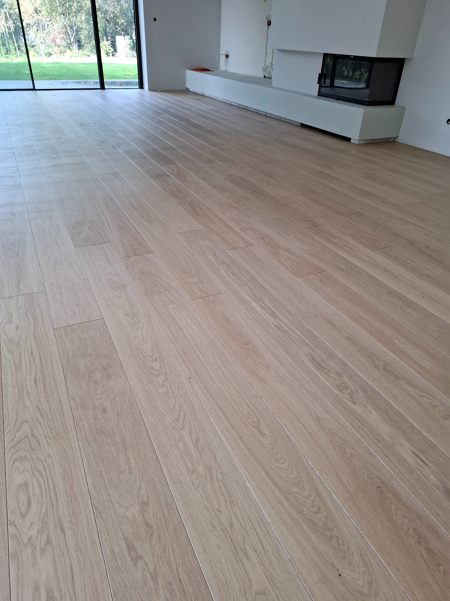 Jasny parkiet ułożony w nowym, minimalistycznym salonie z kominkiem i dużym oknem z widokiem na zieleń.