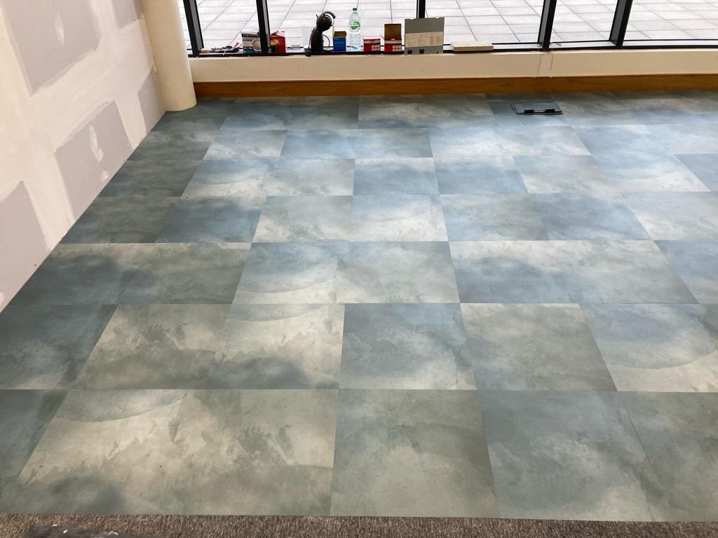 Świeżo położona podłoga z kwadratowych płytek w odcieniach szarości i błękitu, imitujących marmur, widoczna przy ścianie z częściowo wykończonymi płytami gipsowo-kartonowymi i oknem z widokiem...