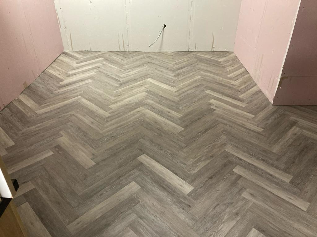 Ukończona podłoga z szarych paneli ułożonych we wzór jodełki w surowym wnętrzu z niepomalowanymi ścianami z płyt gipsowo-kartonowych.