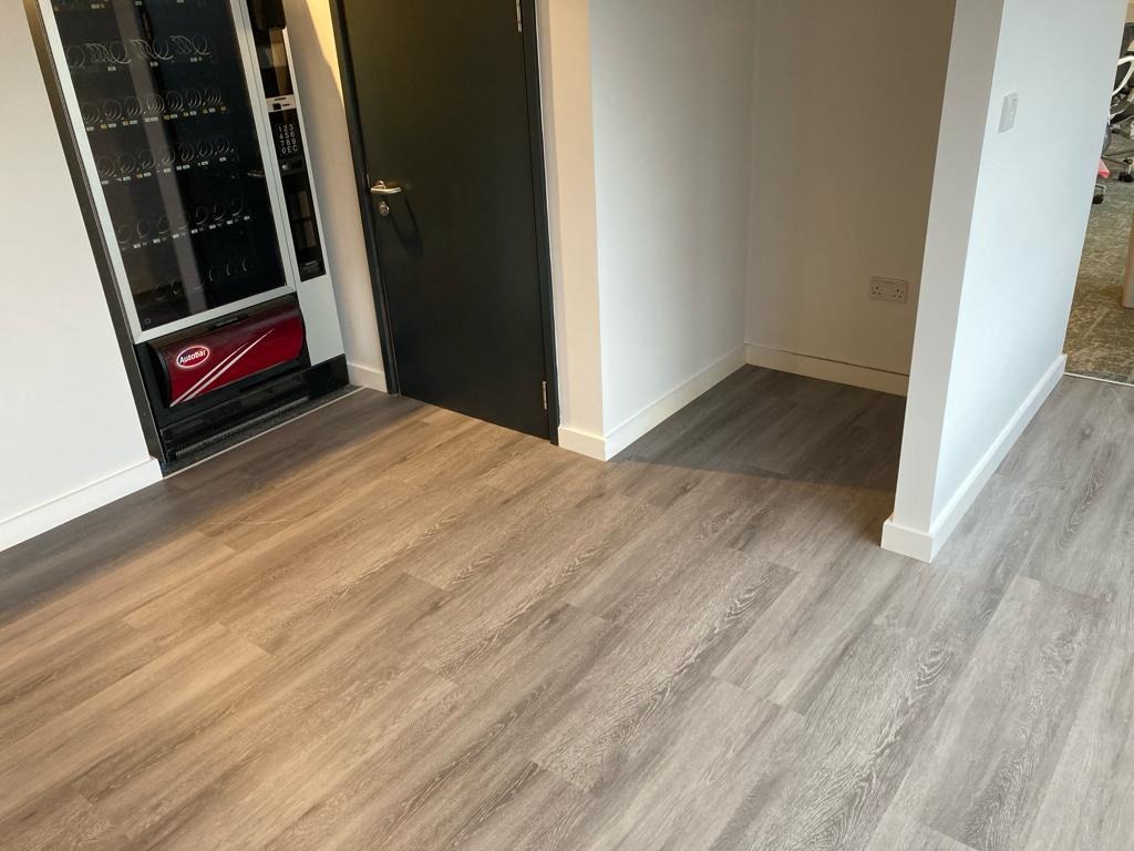 Nowa, jasna podłoga z paneli imitujących drewno w biurze, obok automatu z napojami i czarnych drzwi.