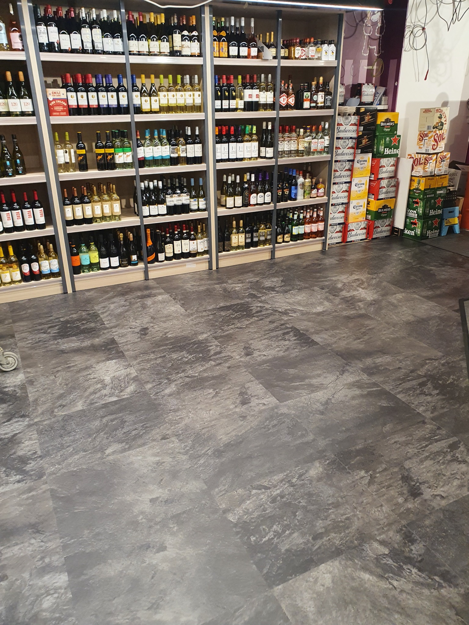 Sklep z alkoholem, widok na szare panele podłogowe imitujące kamień, regały z winami i piwami w tle.