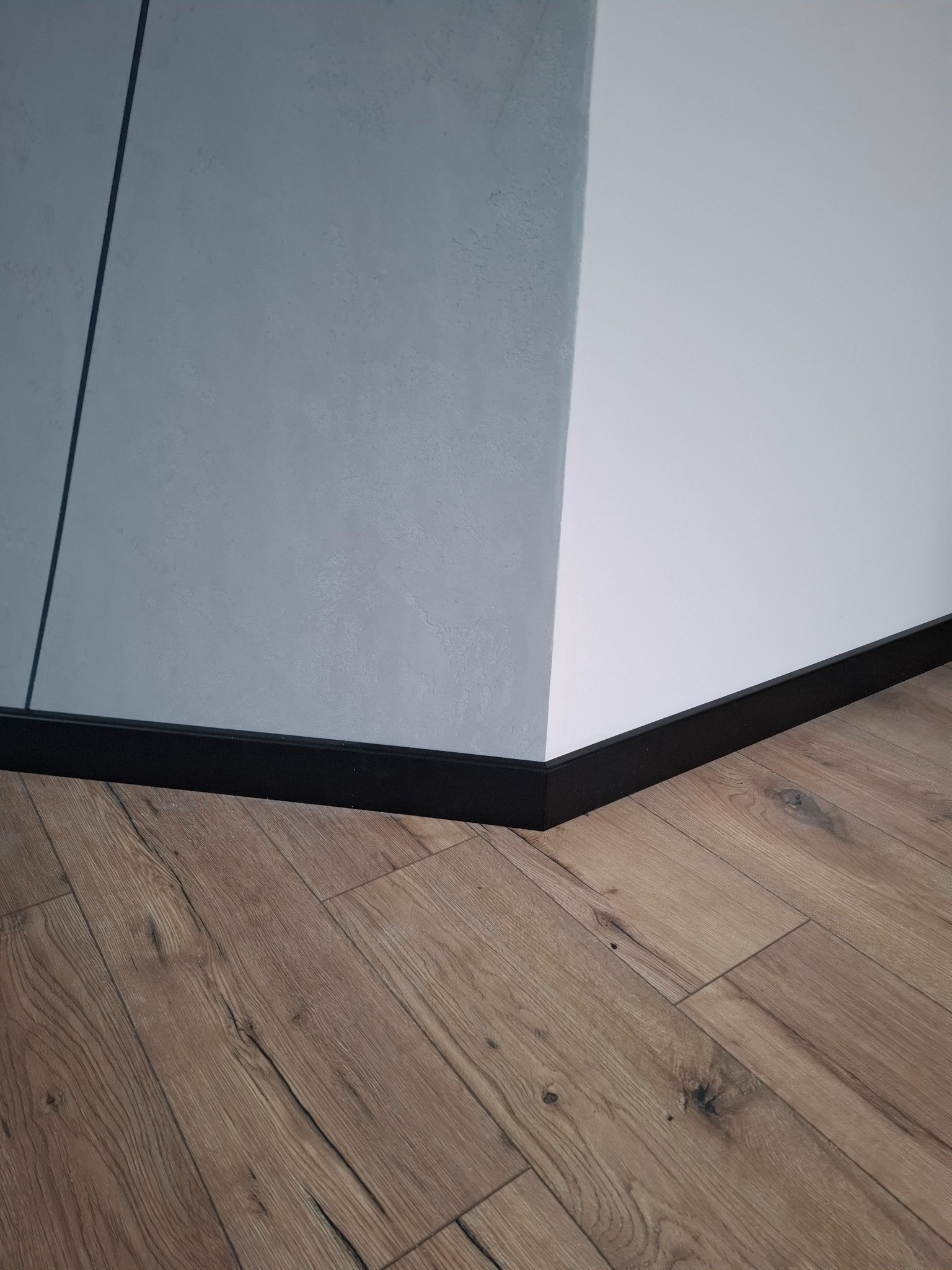 Wykończony róg pomieszczenia z panelami podłogowymi w odcieniach brązu i czarną listwą przypodłogową przy ścianach w kolorze białym i szarym.