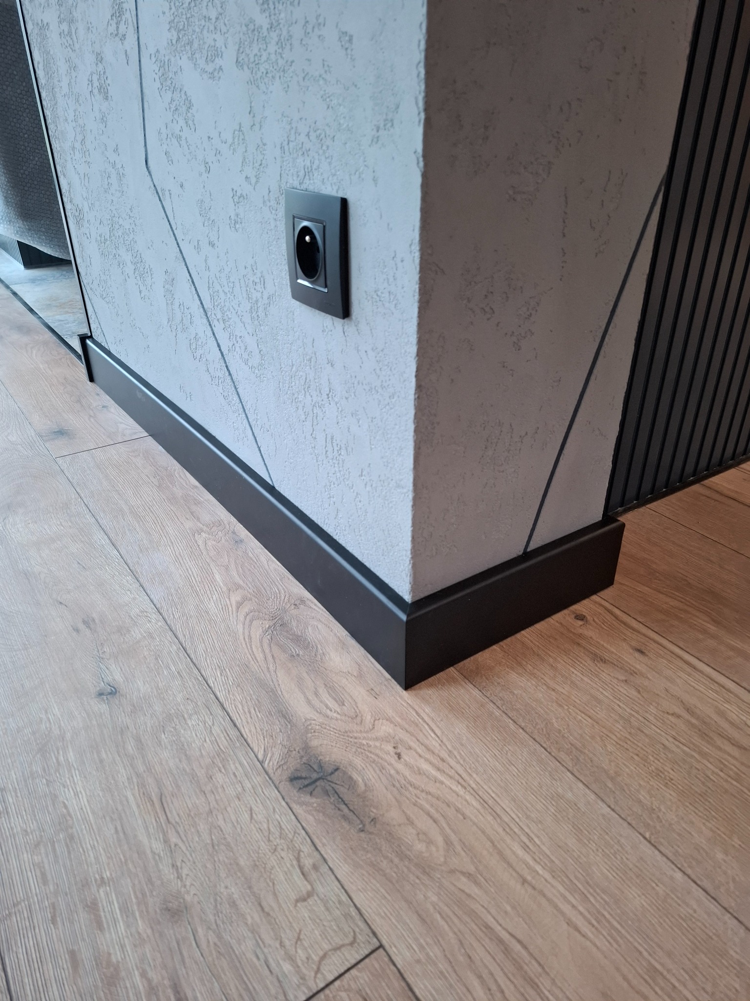 Jasne panele podłogowe z widocznymi słojami drewna, czarna listwa przypodłogowa, szara ściana z dekoracyjną fakturą i czarnym gniazdkiem elektrycznym.