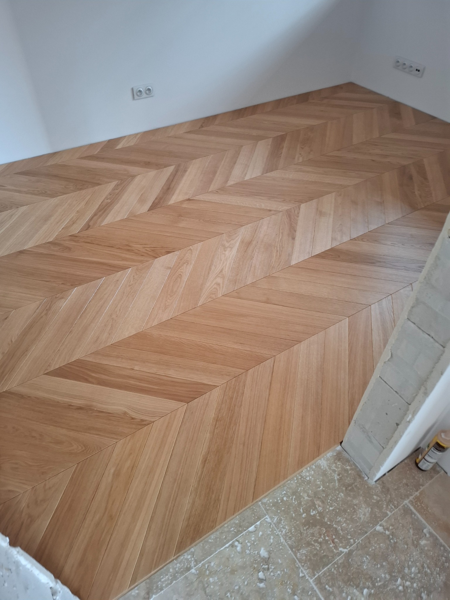 Parkiet ułożony we wzór jodełki francuskiej, widoczne gniazdka elektryczne i fragment surowego wykończenia ściany.