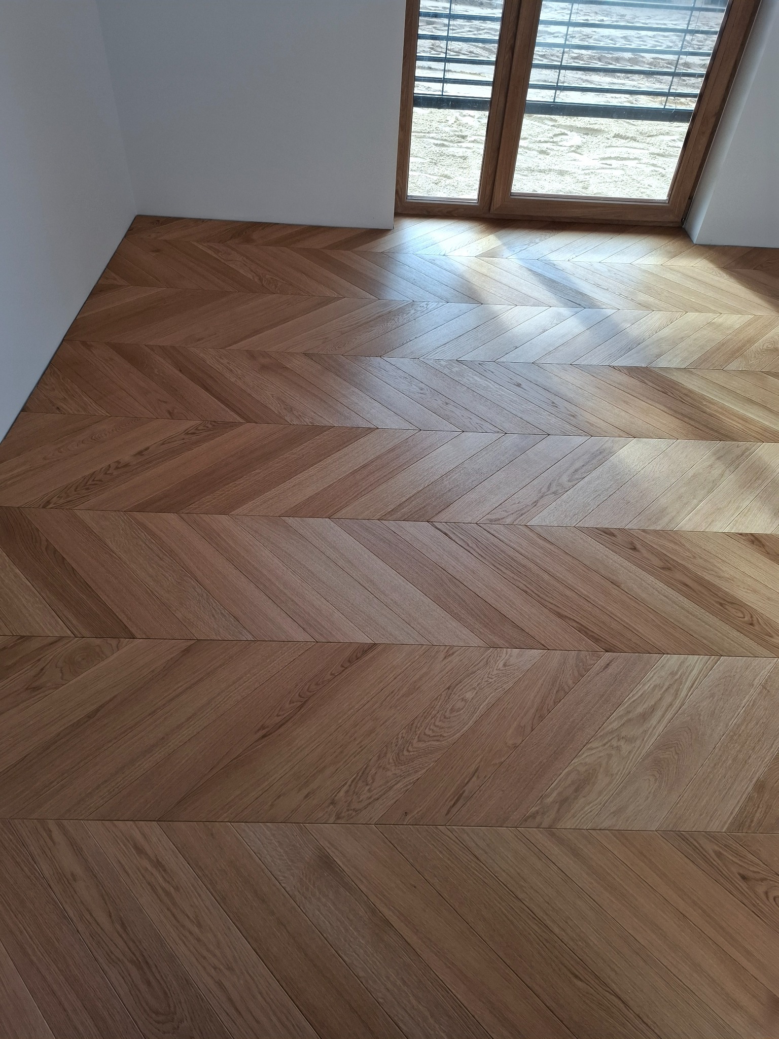 Ułożony parkiet w jodełkę francuską z naturalnego drewna, widok na okno z żaluzjami i jasny piasek za nim.