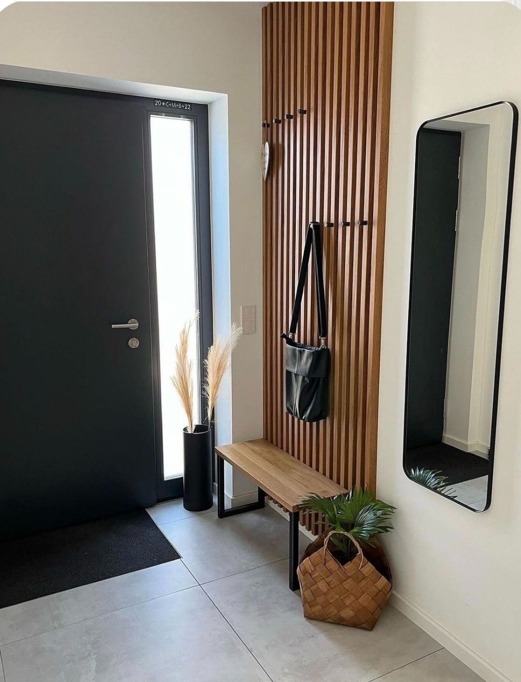 Nowoczesny przedpokój z szarą podłogą, czarnymi drzwiami wejściowymi z szybą, drewnianym panelem ściennym z wieszakami, lustrem w czarnej ramie, drewnianą ławką z czarnymi nogami, czarnym wazonem...