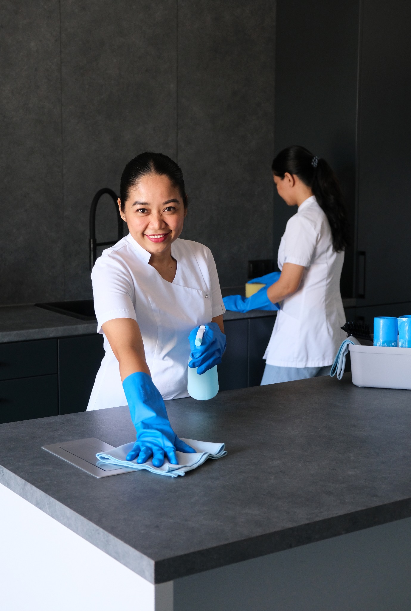 Uśmiechnięta sprzątaczka w niebieskich rękawicach przeciera szarą powierzchnię blatu kuchennego, w tle druga osoba z gąbką i pojemnik z detergentami.