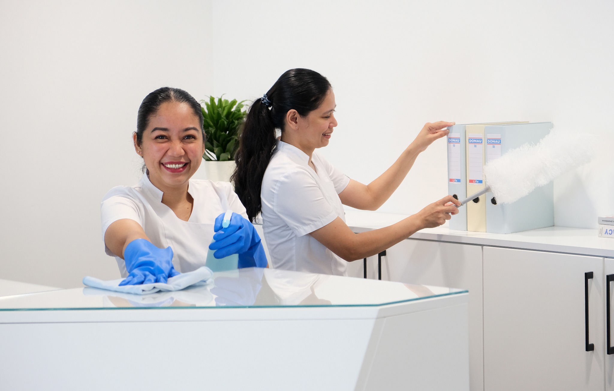 Dwie uśmiechnięte kobiety w białych uniformach czyszczą białe biuro; jedna z niebieskimi rękawiczkami przeciera blat, druga odkurza segregatory z dokumentami.