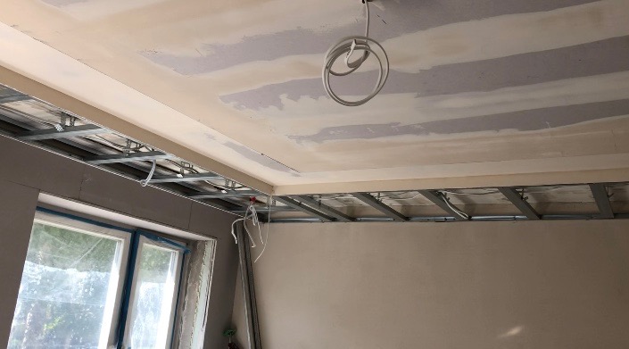 Sufit podwieszany w trakcie montażu: widoczne metalowe profile, częściowo położona płyta gipsowo-kartonowa z widocznymi łączeniami, wiszące przewody elektryczne.