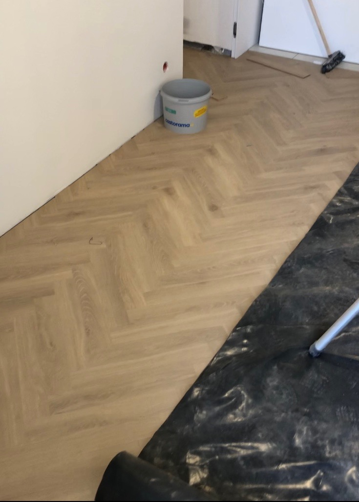 Ułożone panele w jodełkę, częściowo przykryte folią ochronną, widoczne wiadro z farbą Castorama przy ścianie.