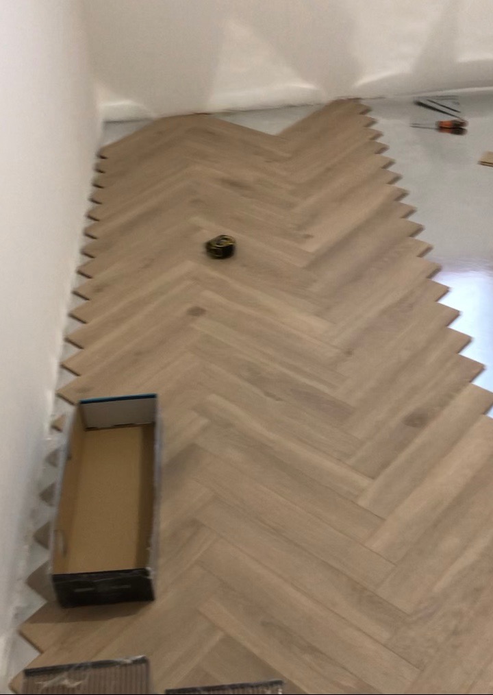 Układanie paneli podłogowych w jodełkę, widoczny etap prac z niedociętymi panelami przy ścianie i pudełko po panelach w rogu pomieszczenia.