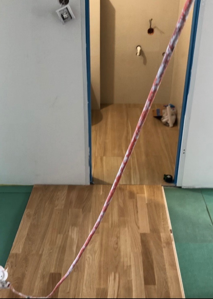 Fragment układania jasnych paneli podłogowych, widoczny sznur traserski i zielona folia ochronna, w tle wykończone pomieszczenie z instalacją elektryczną.