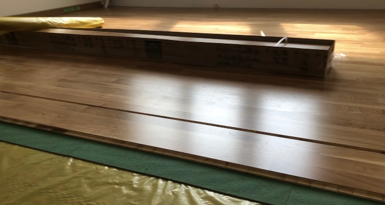 Fragment podłogi z dębowych paneli ułożonych na zielonej macie izolacyjnej, z widocznym opakowaniem paneli w tle, częściowo przykrytym folią ochronną.