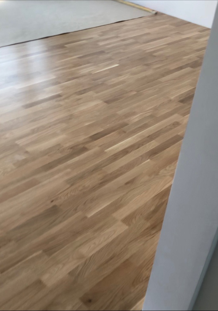 Ułożone panele podłogowe z jasnego drewna, widoczne łączenia desek, fragment ściany w tle.