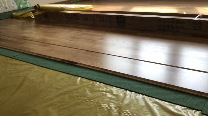 Układanie paneli podłogowych: widoczne panele w trakcie montażu, folia i podkład wygłuszający, fragment rolki folii w tle.