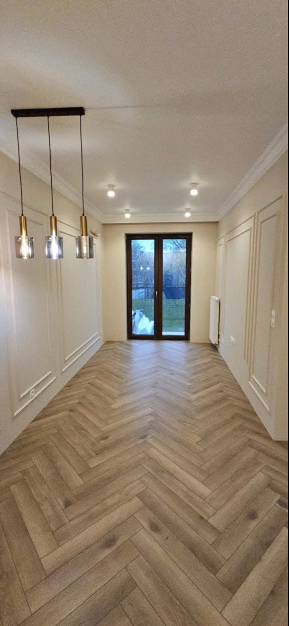 Salon po remoncie: parkiet ułożony w jodełkę, kremowe ściany z dekoracyjnymi listwami, czarne drzwi balkonowe i nowoczesna lampa wisząca z trzema szklanymi kloszami.