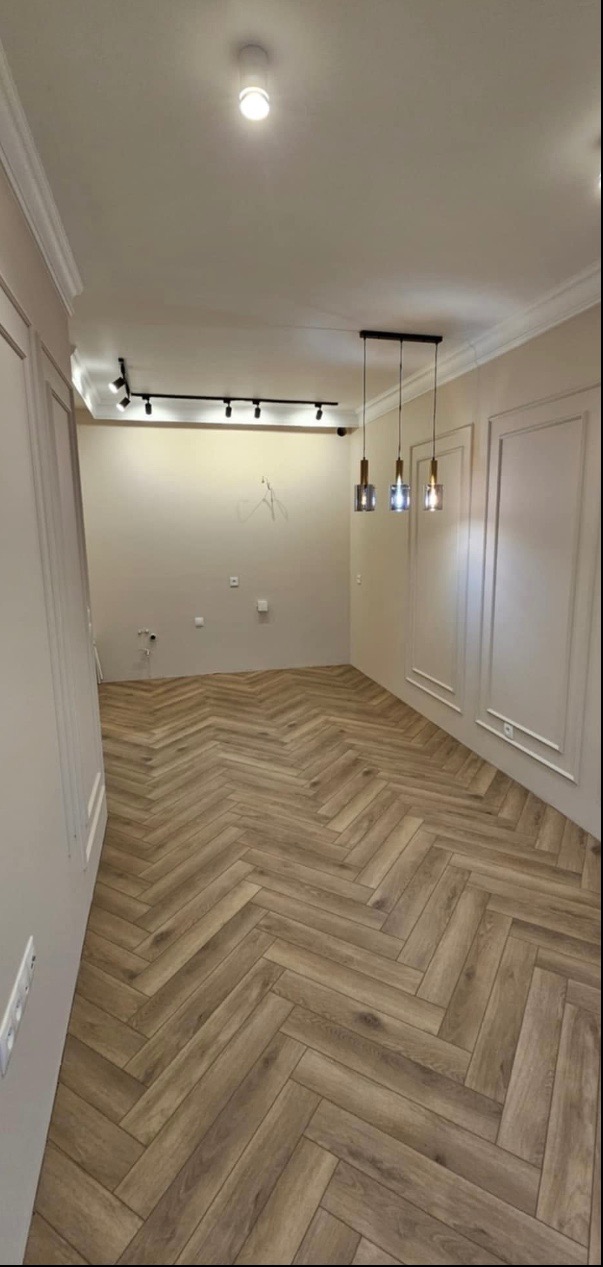 Salon w Róży z ułożonymi panelami w jodełkę, dekoracyjne listwy ścienne, oświetlenie szynowe i wiszące lampy szklane.