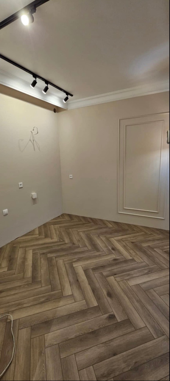 Pokój po remoncie z ułożoną podłogą z paneli w jodełkę, beżowe ściany z ozdobną sztukaterią i nowoczesnym oświetleniem szynowym.