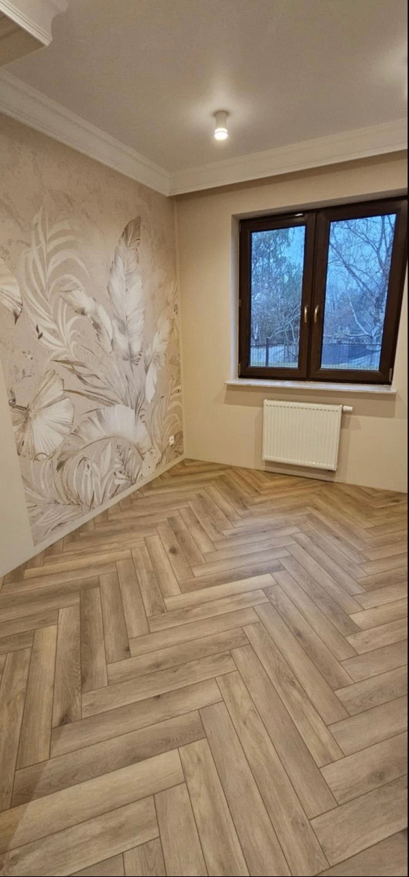 Pokój po remoncie z nową podłogą ułożoną w jodełkę, ścianą z motywem roślinnym i oknem z brązową ramą.