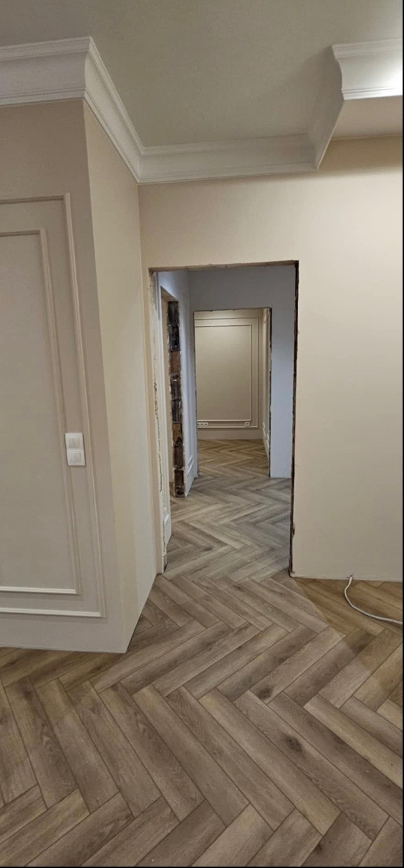 Korytarz po remoncie z panelami ułożonymi w jodełkę, widoczne listwy przypodłogowe i drzwi z ozdobnymi ramami, w tle kolejne pomieszczenia z podobnym wykończeniem.