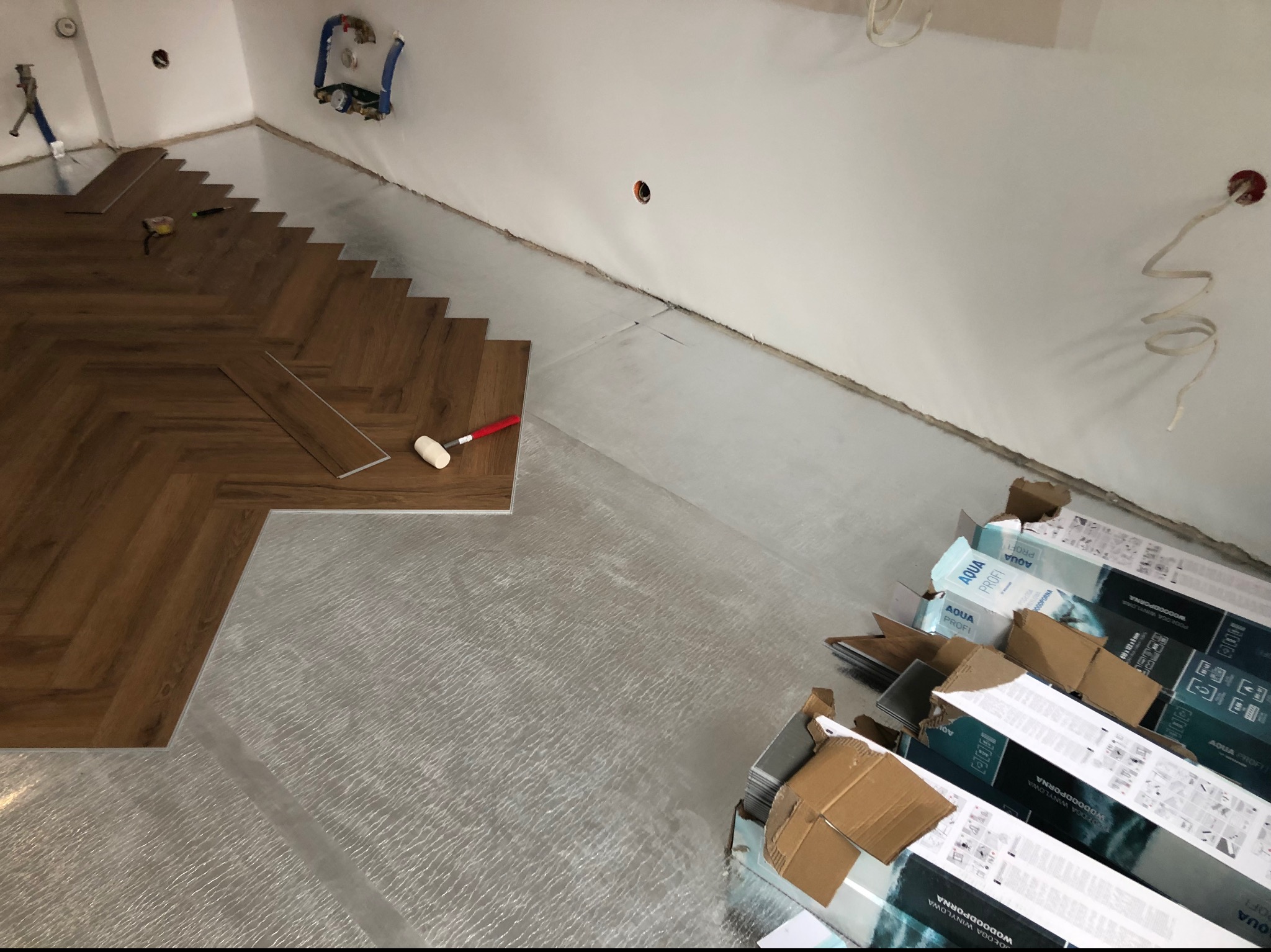 Układanie paneli w jodełkę na podkładzie izolacyjnym, widoczne narzędzia i opakowania paneli Aqua Profi.