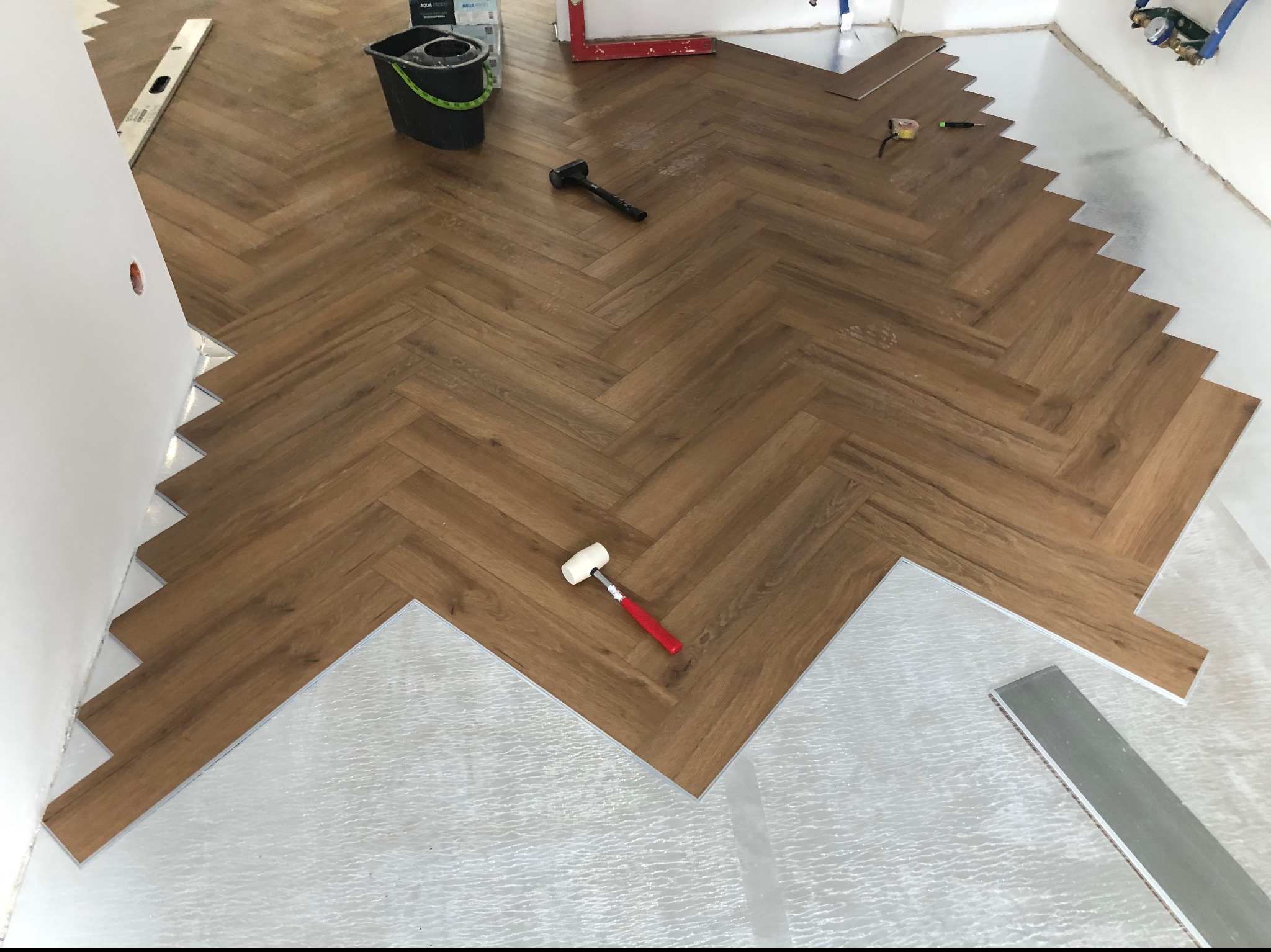 Układanie paneli podłogowych w jodełkę, widoczna izolacja podkładowa, gumowy młotek z czerwoną rączką i czarny młotek ślusarski na powierzchni.