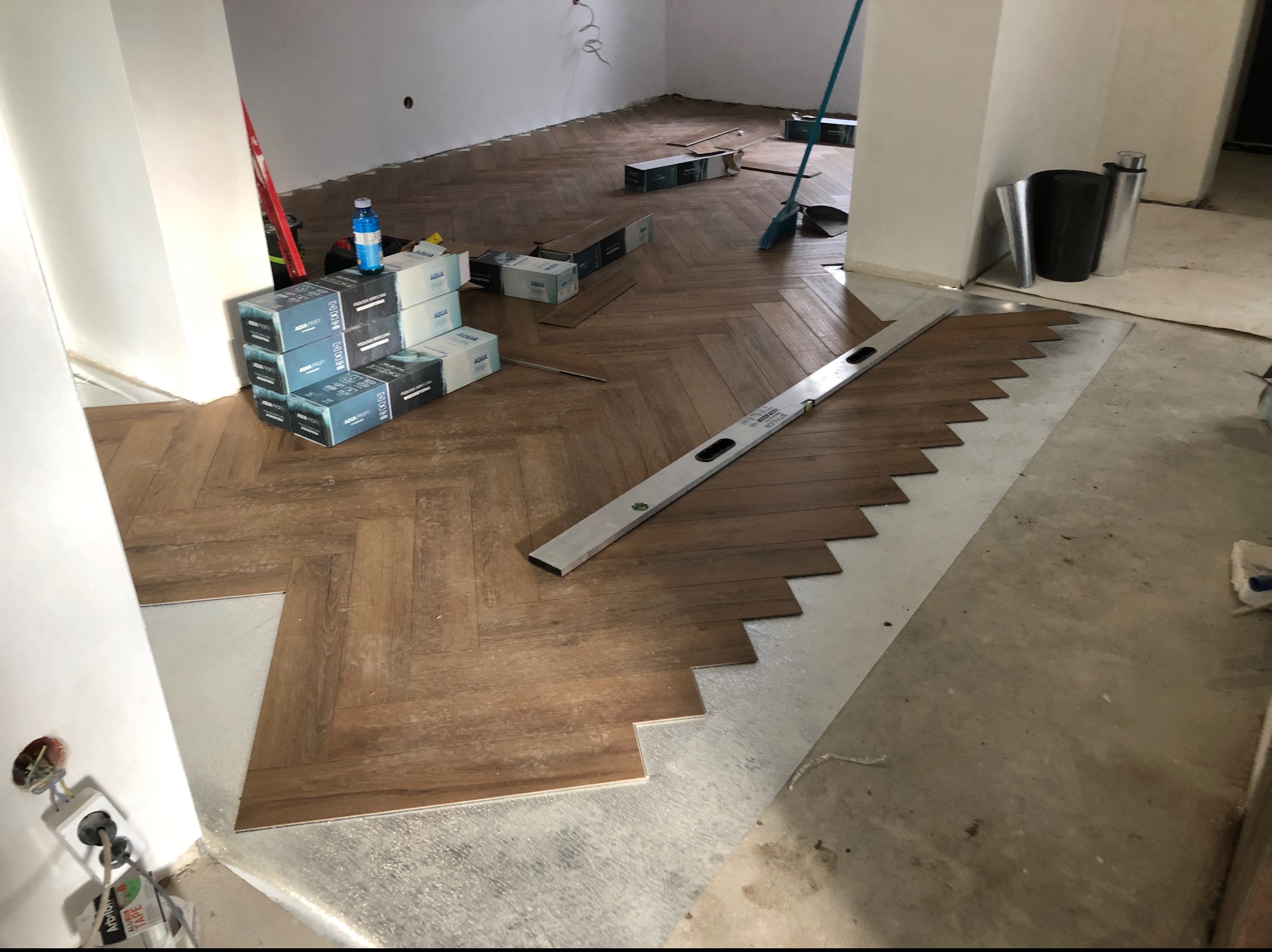 Układanie paneli podłogowych w jodełkę, widoczny poziomica, paczki paneli Aqua Profi oraz fragmenty podkładu pod panele.