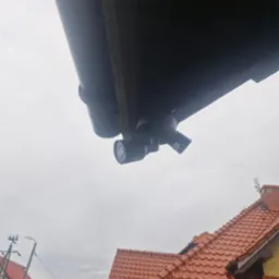 Zewnętrzne kamery monitoringu zamontowane pod dachem domu, widok z dołu na tle pochmurnego nieba i fragmentu dachu z czerwoną dachówką.