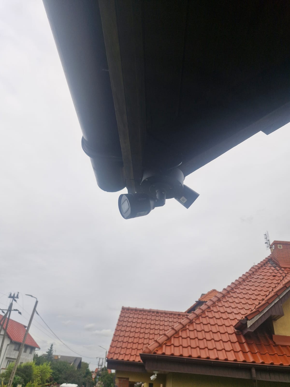 Zewnętrzne kamery monitoringu zamontowane pod dachem domu, widok z dołu na tle pochmurnego nieba i fragmentu dachu z czerwoną dachówką.