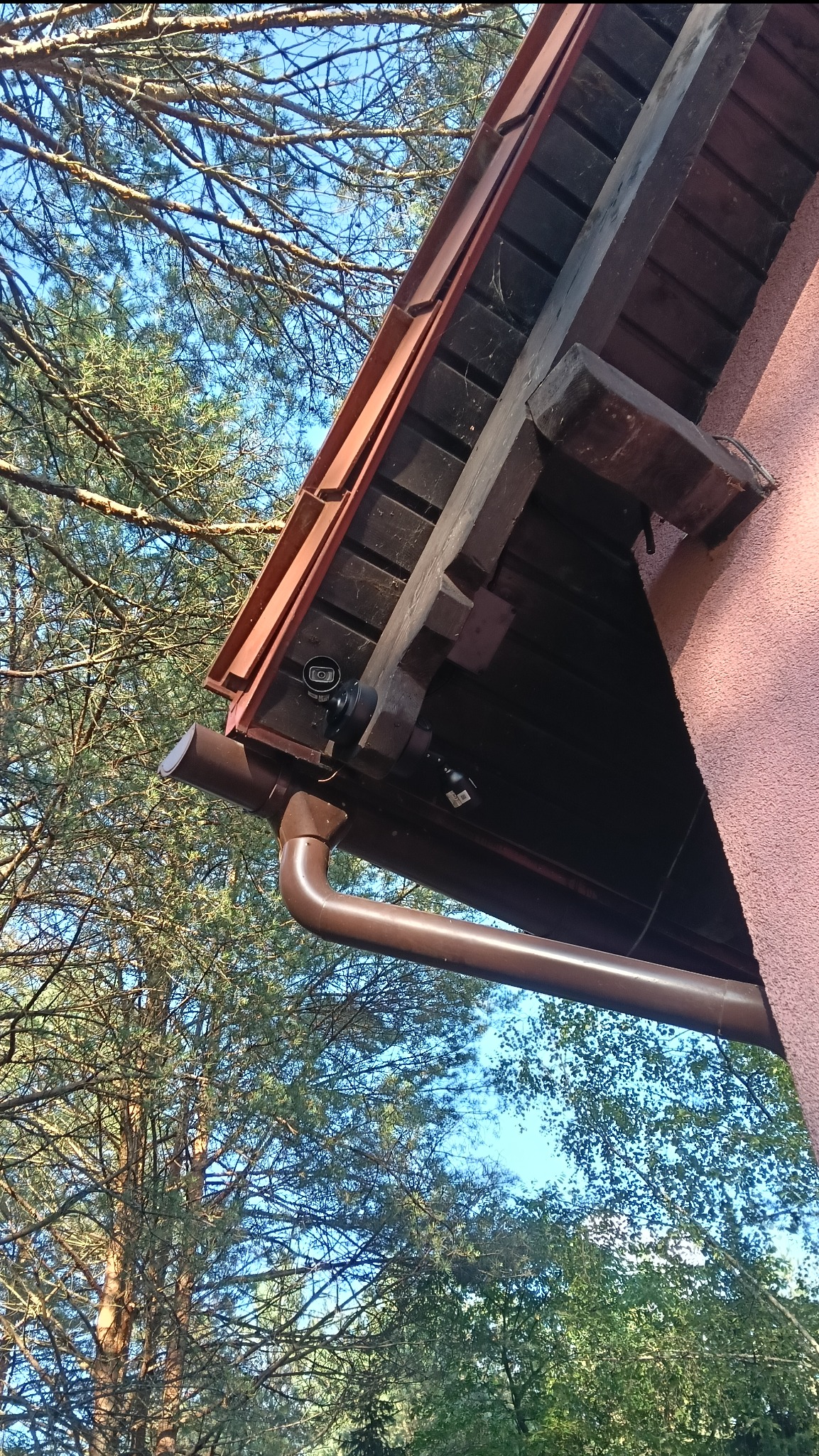 Kamera monitoringu zamontowana pod okapem drewnianego domu, widok z dołu na tle błękitnego nieba i zieleni drzew. Brązowa rynna.