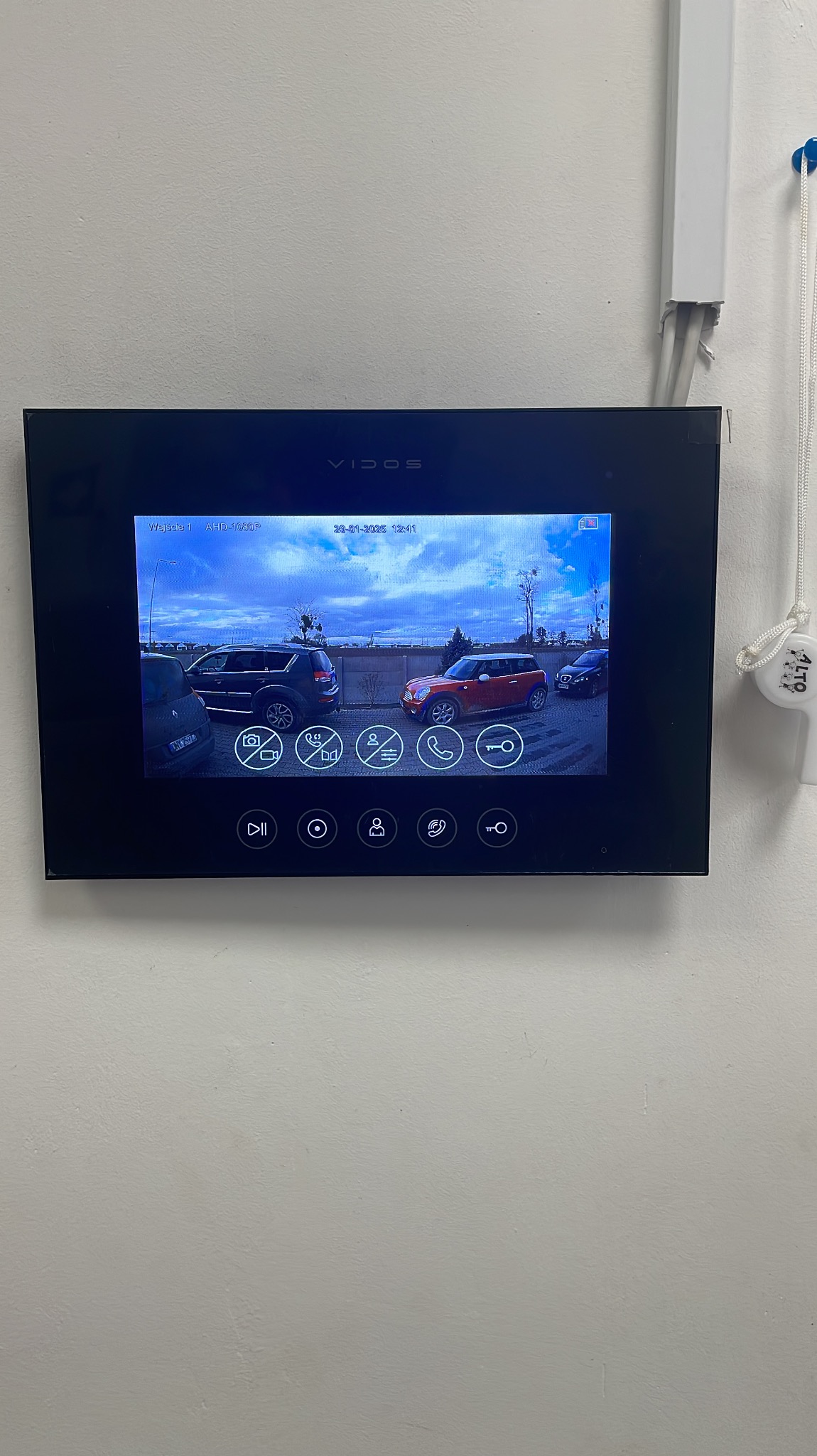 Zamontowany na ścianie panel wideodomofonu marki Vidos, wyświetlający obraz z kamery z widokiem na podjazd z zaparkowanymi samochodami w pochmurny dzień.
