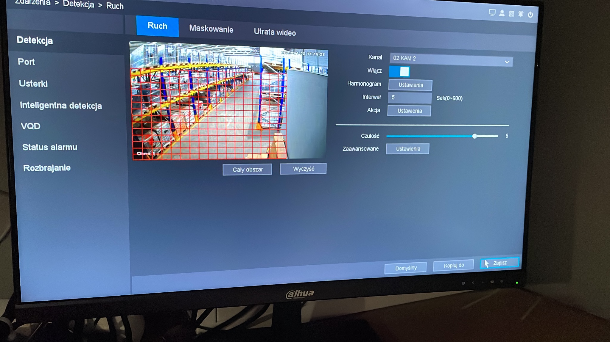 Ekran monitora z ustawieniami detekcji ruchu kamery przemysłowej w magazynie z regałami, widoczne opcje konfiguracji i podgląd z kamery z naniesioną siatką detekcji.