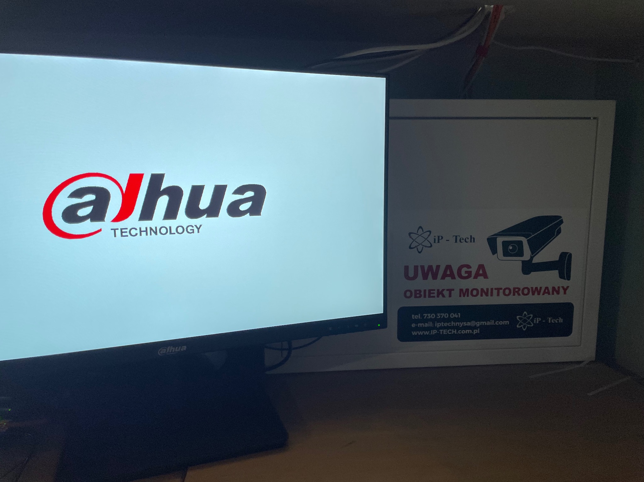 Monitor z wyświetlonym logo Dahua Technology, obok tabliczka 'Uwaga Obiekt Monitorowany' z logiem iP-Tech, wskazująca na system monitoringu.