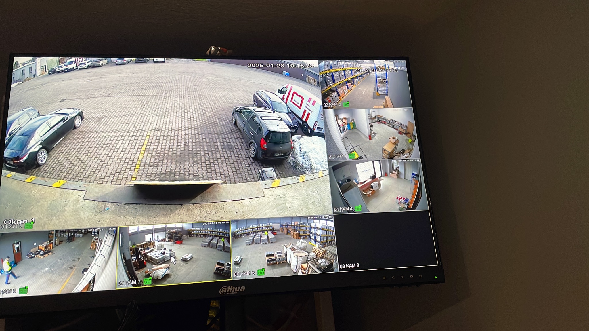 Ekran monitora z podglądem z kamer przemysłowych obejmujących magazyn, plac manewrowy i wnętrze biura, data i numer kamery w rogu każdego okna podglądu.