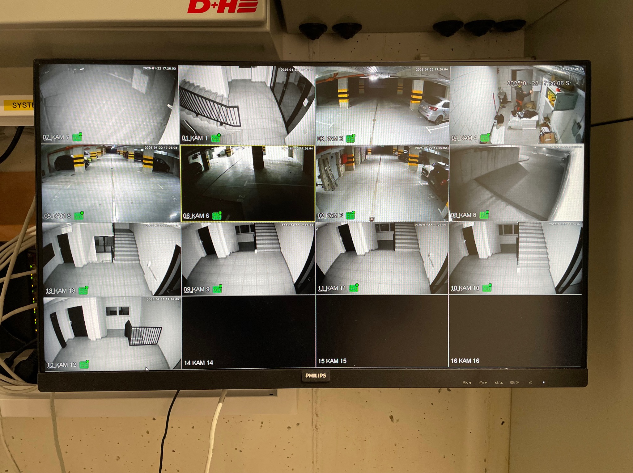 Monitor Philips wyświetlający obraz z szesnastu kamer monitoringu, pokazujących różne obszary, w tym parking podziemny i klatkę schodową.