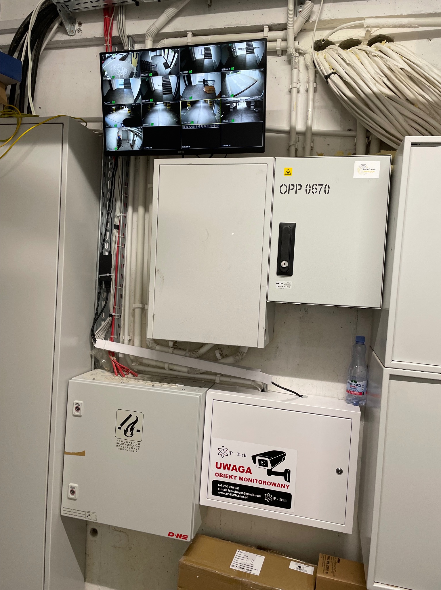 Centrum sterowania systemem monitoringu wizyjnego: monitor z podglądem z kamer, szafy sterownicze, okablowanie, etykieta 'Obiekt Monitorowany' na szafce.
