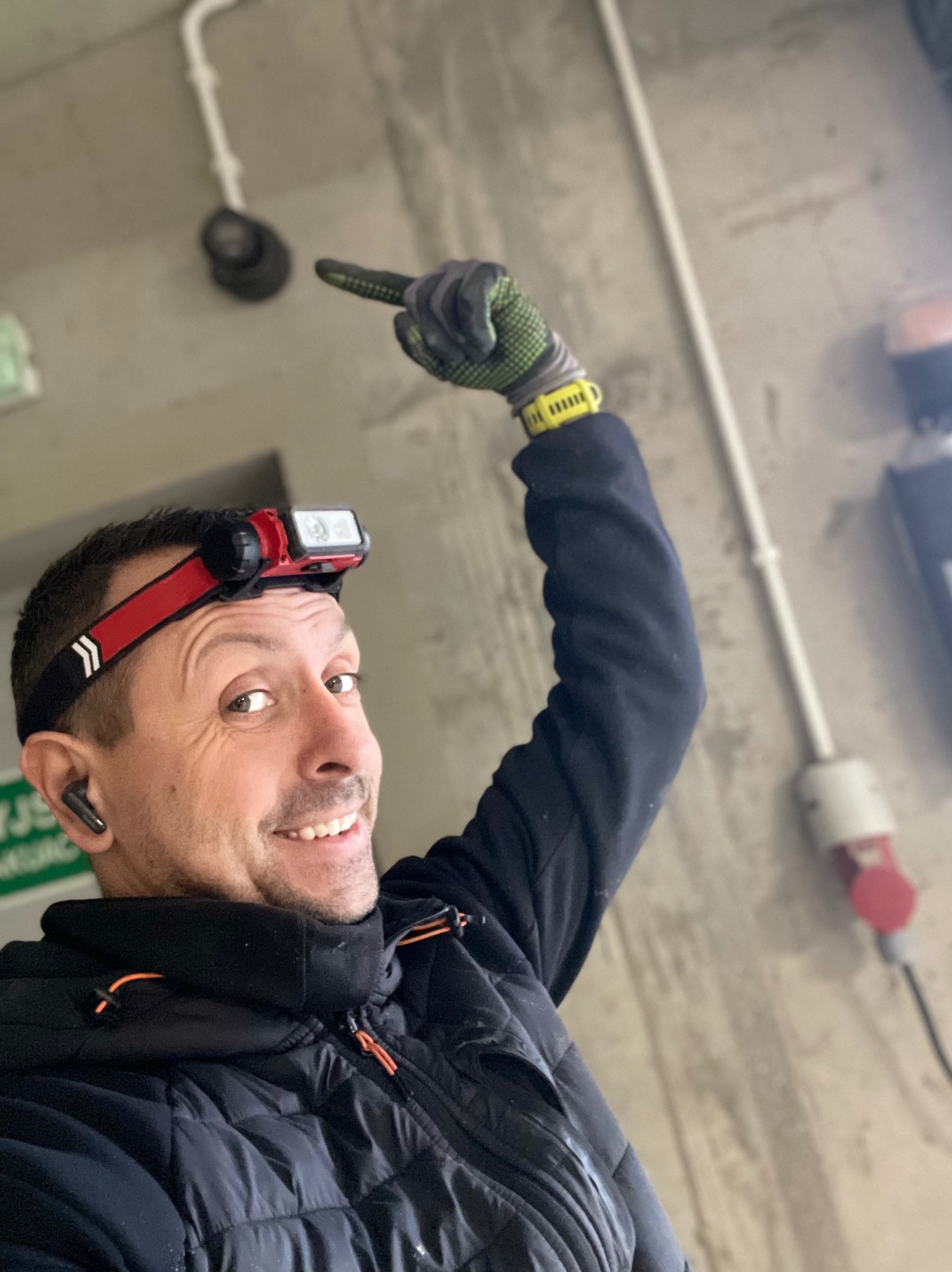 Uśmiechnięty elektryk z czołówką, w kurtce i rękawicach ochronnych, palcem wskazującym na czujnik dymu zamontowany na suficie. Widoczna instalacja elektryczna i gniazdo siłowe w tle.