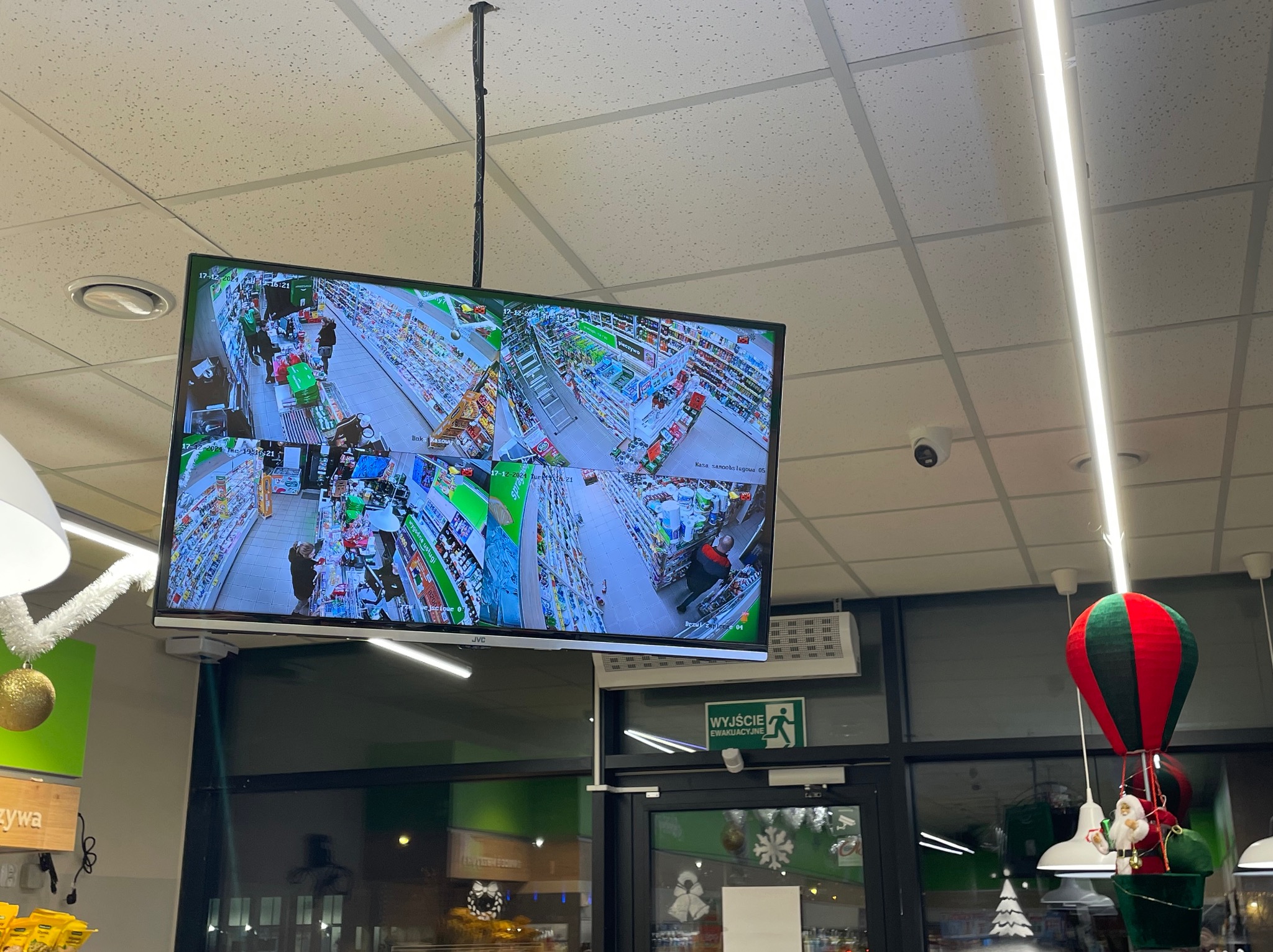 Ekran monitoringu w sklepie spożywczym, wyświetlający obraz z kilku kamer CCTV, zawieszony pod sufitem, widoczny również czujnik ruchu i oświetlenie.