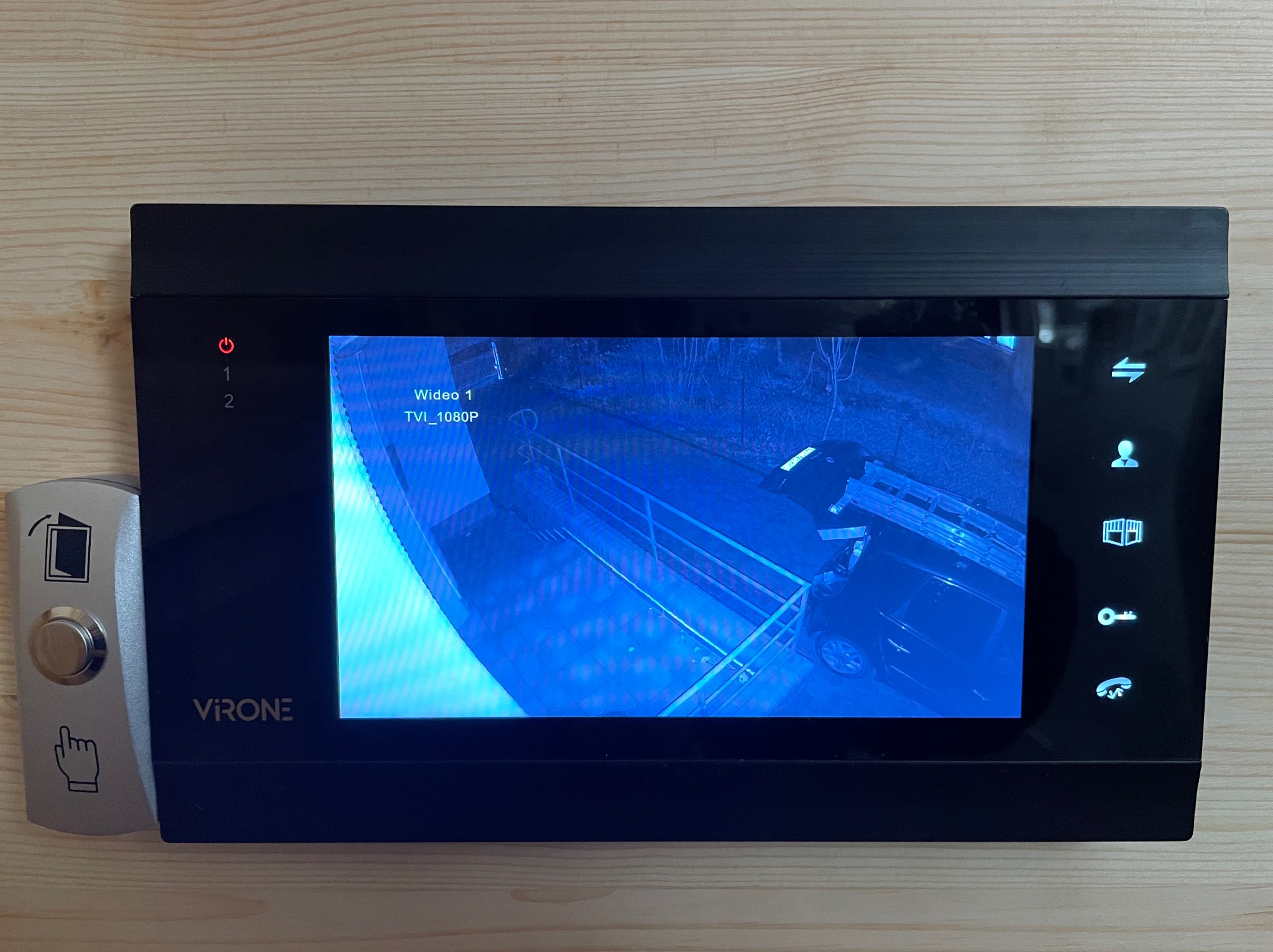 Czarny wideodomofon VIRON z widokiem na zewnątrz, ukazujący fragment podjazdu z samochodem i drabiną opartą o budynek, zamontowany na drewnianej ścianie obok srebrnego przycisku dzwonka z symbolem...