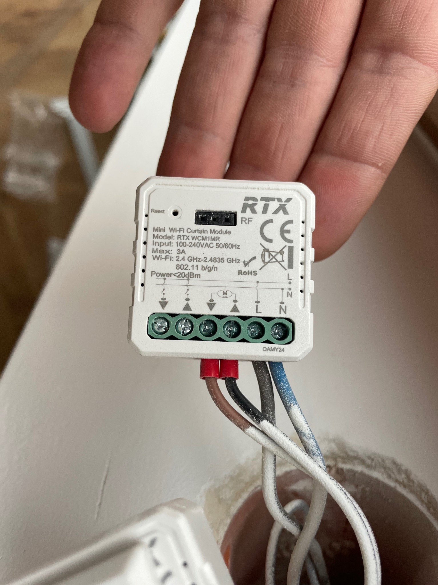 Miniaturowy moduł Wi-Fi do sterowania zasłonami RTX WCM1MR, trzymany w dłoni, z podłączonymi przewodami, widoczne oznaczenia L i N na module.