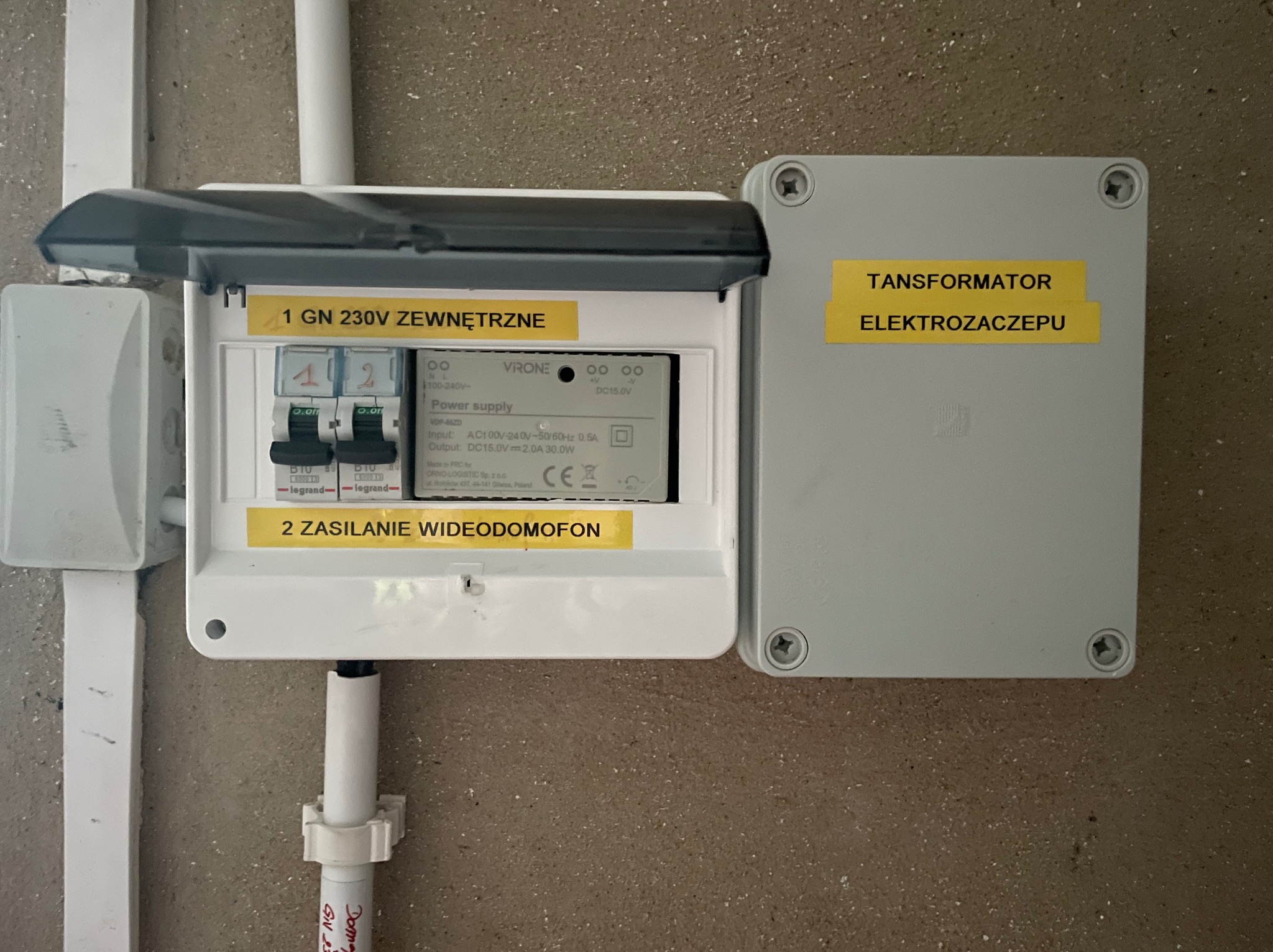 Rozdzielnica elektryczna zasilająca wideodomofon i inne obwody, z otwartą obudową i widocznymi bezpiecznikami oraz transformatorem elektro zaczepu obok.