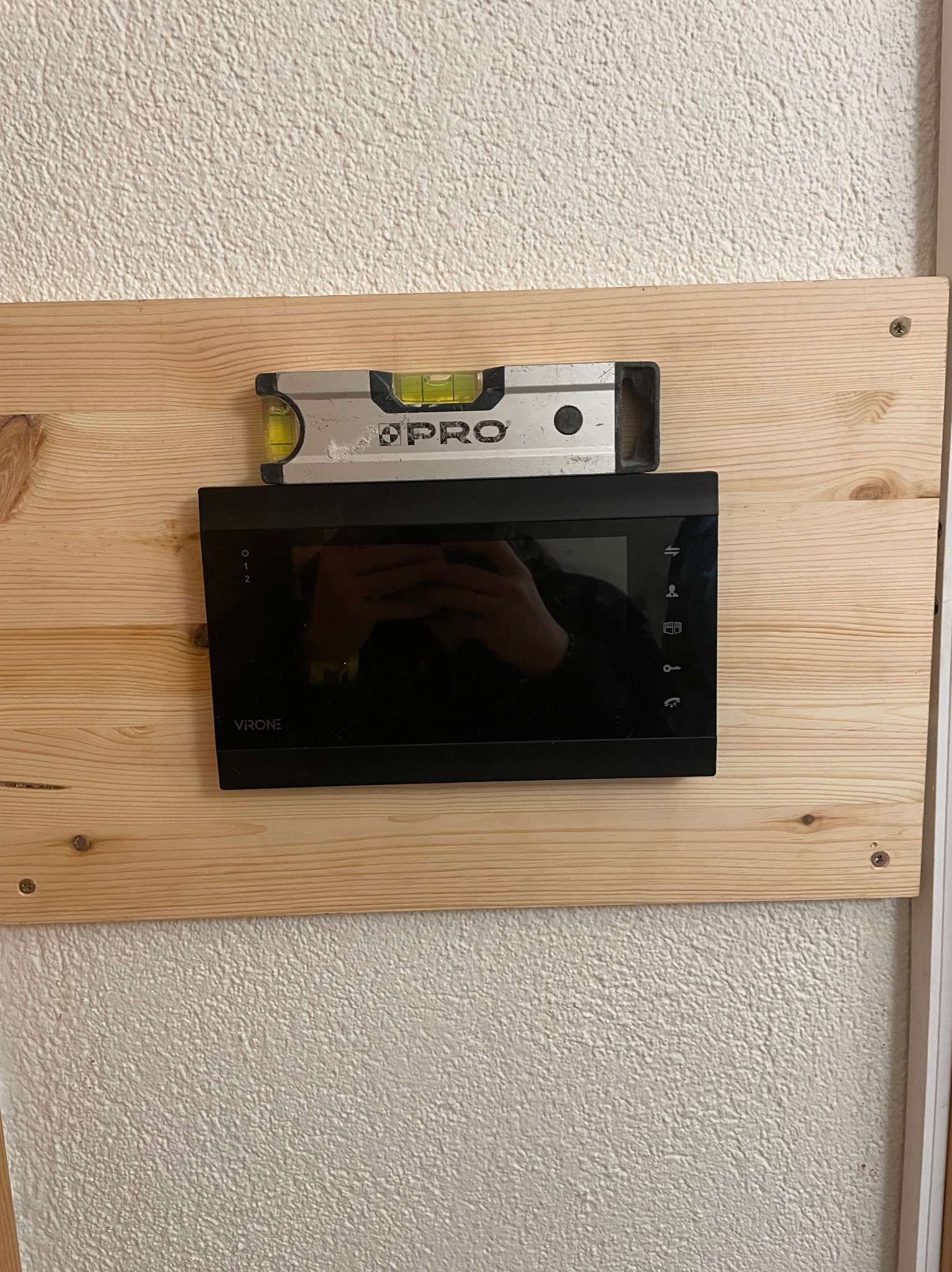 Czarny wideofon Vrone zamontowany na drewnianej płycie, poziomica PRO u góry, widoczna faktura ściany.