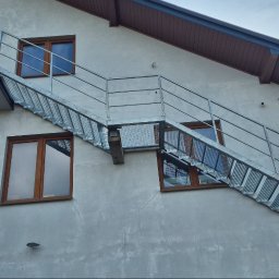 LEKSTAL KONSTRUKCJE - Zewnętrzne schody metalowe z ażurowymi stopniami, prowadzące do balkonu na elewacji budynku z drewnianymi oknami. Widok z dołu, pochmurne niebo w tle.