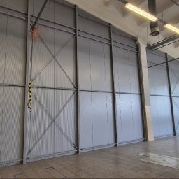 LEKSTAL KONSTRUKCJE - Wnętrze hali z szarymi płytami warstwowymi i metalową konstrukcją. Widoczne oświetlenie sufitowe i okno. Na podłodze leżą narzędzia.