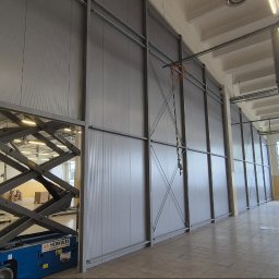 LEKSTAL KONSTRUKCJE - Wnętrze hali w trakcie montażu płyt warstwowych, widoczny podnośnik nożycowy i częściowo zamontowana ściana z płyt. Oświetlenie przemysłowe, posadzka z płytek.