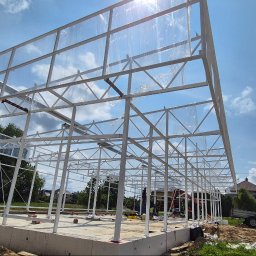 LEKSTAL KONSTRUKCJE - Konstrukcja stalowa hali w trakcie budowy, widok z zewnątrz, białe profile, częściowo pokryte folią, fundament betonowy, błękitne niebo w tle.